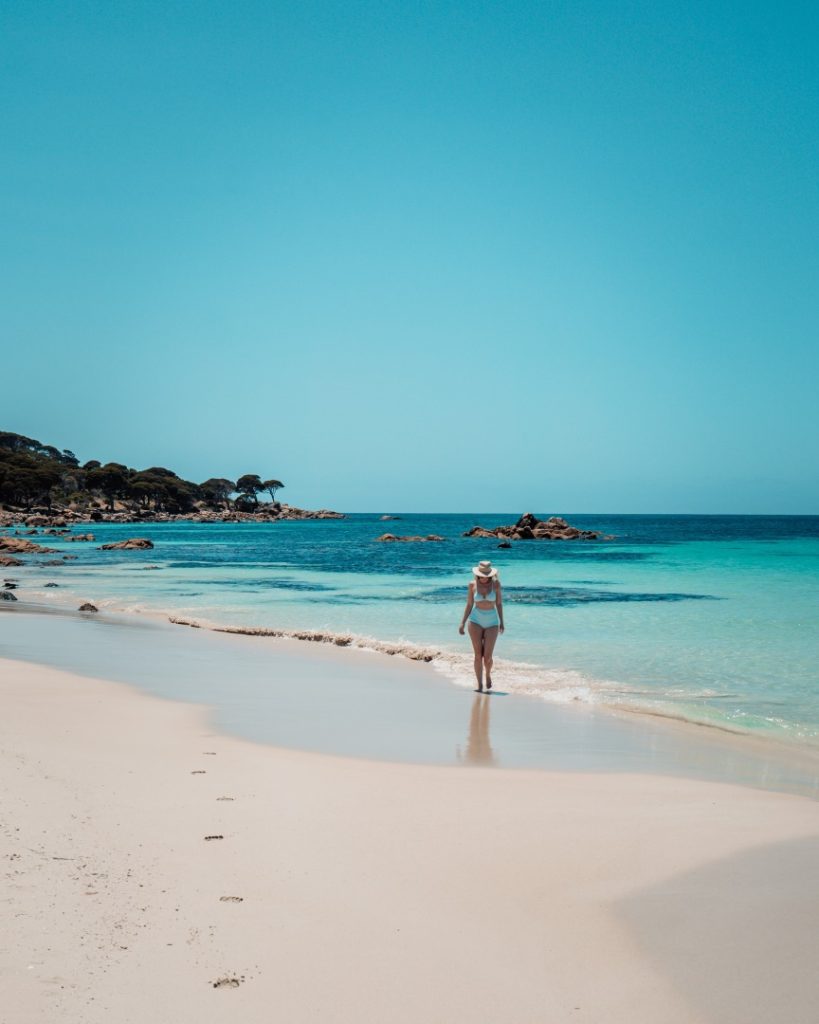 A woman walks along the pristine sandy shore of Bunker Bay Beach in Western Australia, with clear blue waters and rocky outcrops in the background.