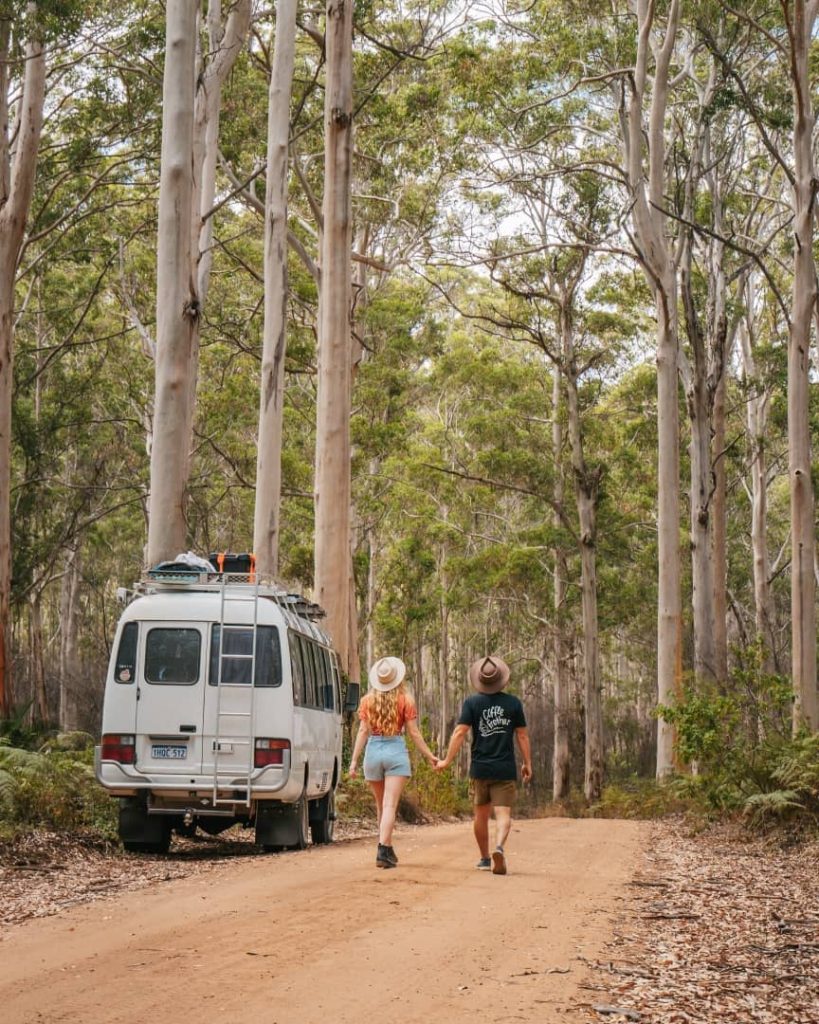 A couple walks hand in hand along a dirt road flanked by tall trees in Boranup Drive, Margaret River, Western Australia. A white camper van is parked nearby.