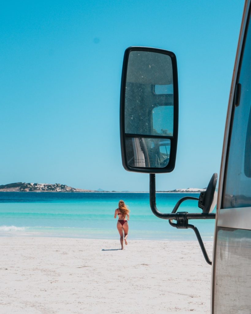 A woman walking on the white sands of a secluded Esperance beach, captured through the side mirror of a campervan.