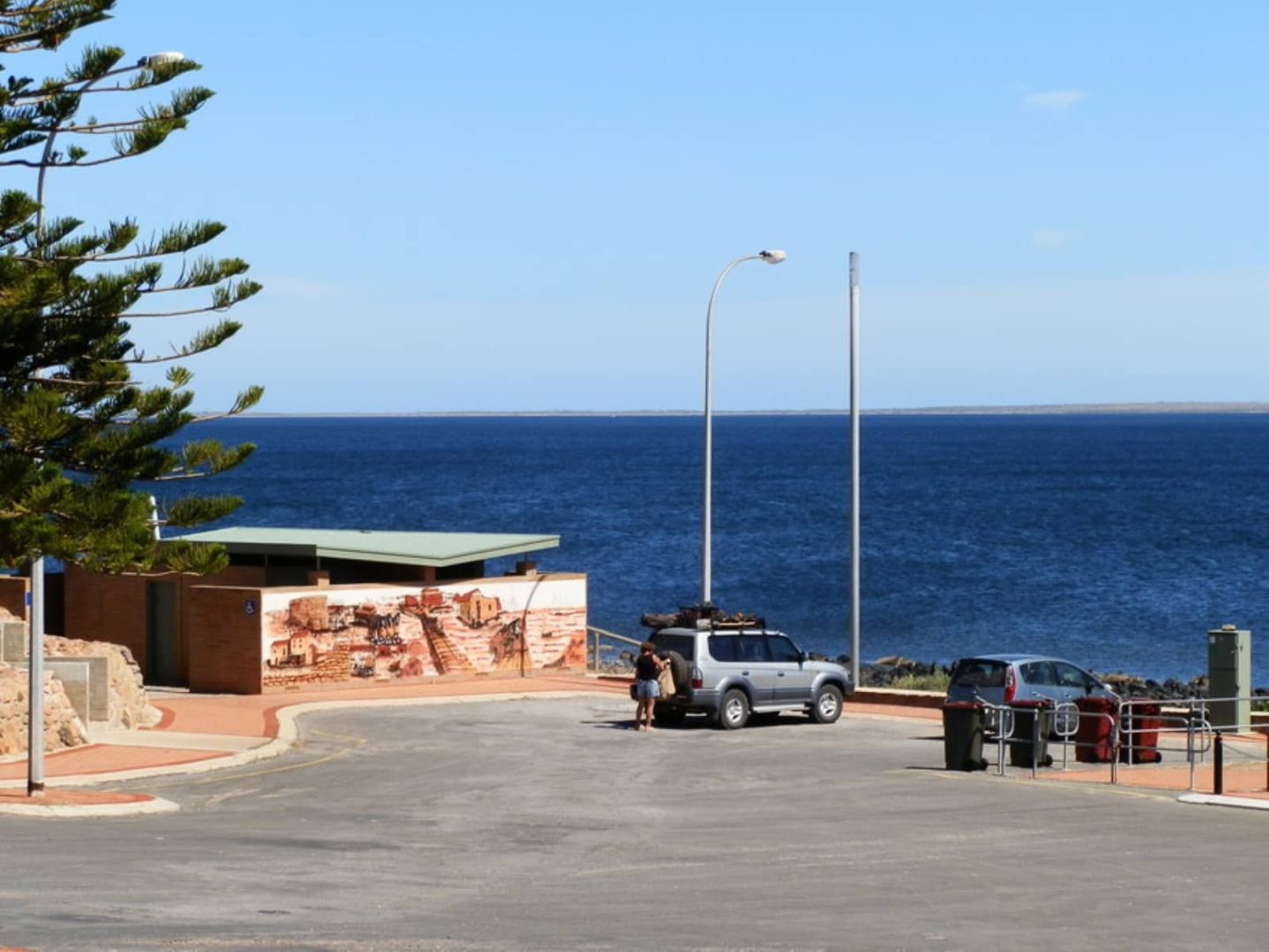 View of the ocean and mural-decorated public toilets at the Port Victoria foreshore car park, with a couple standing near a 4WD on a Yorke Peninsula road trip.