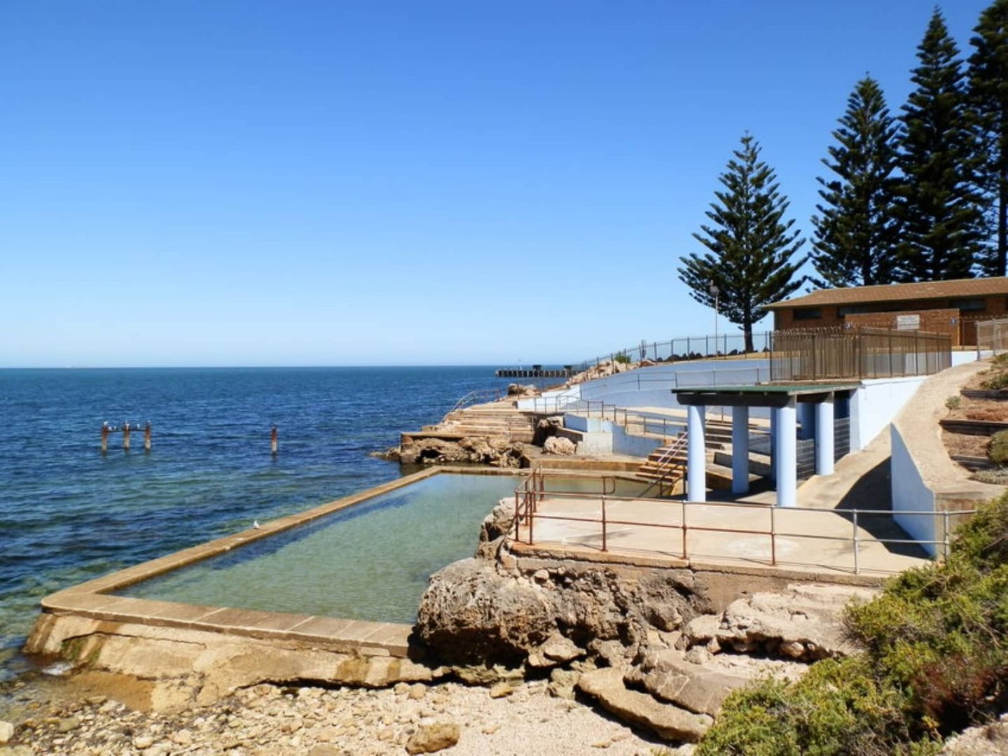 Seawater swimming pool built into the rocky coast beside the ocean, bordered by railings and pine trees, captured on a sunny Yorke Peninsula road trip.