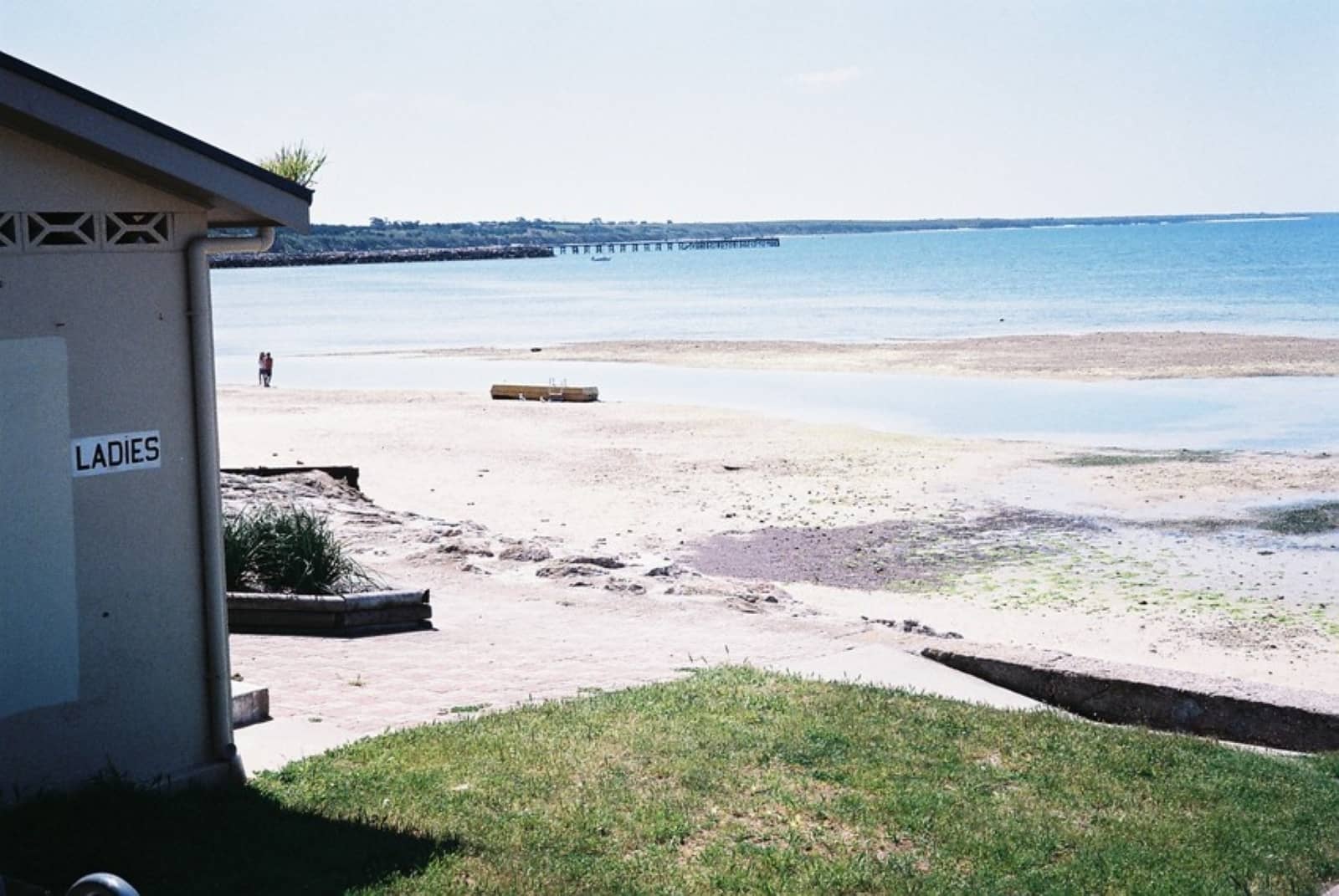 A sandy beach with calm water seen next to a public restroom building marked โLADIES,โ with a couple walking in the distance during a Yorke Peninsula road trip.