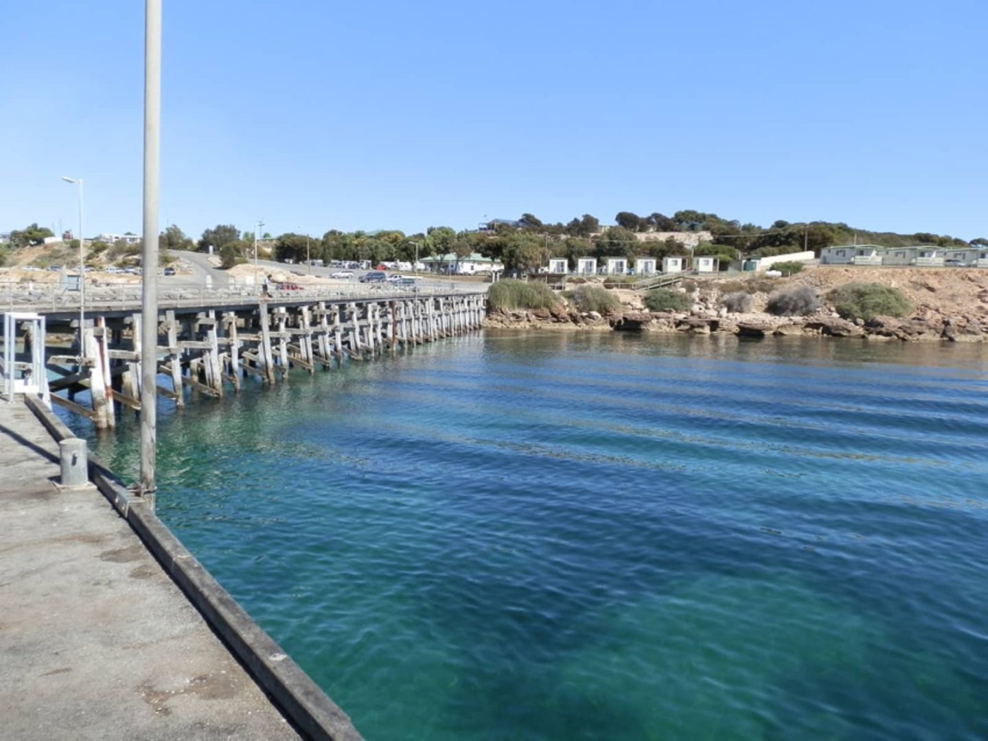 A wooden jetty extending into calm, clear blue water with coastal cabins and rocky shoreline behind it, seen on a Yorke Peninsula road trip.