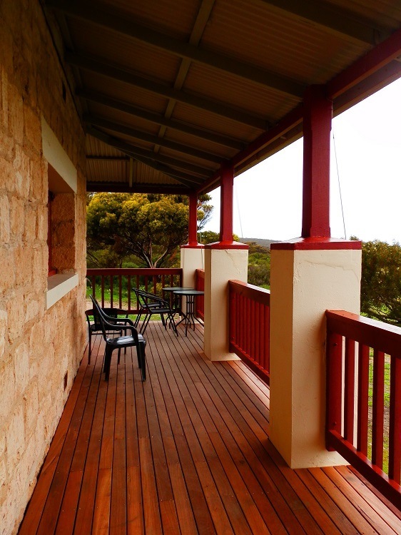 The outside of Engineers House in Innes NP, Yorke Peninsula, SA. Covered wooden veranda attached to a rustic stone cottage, featuring black chairs and small round tables along red railings. The porch overlooks a green, tree-filled landscape under a cloudy sky, offering a quiet, sheltered outdoor seating area.