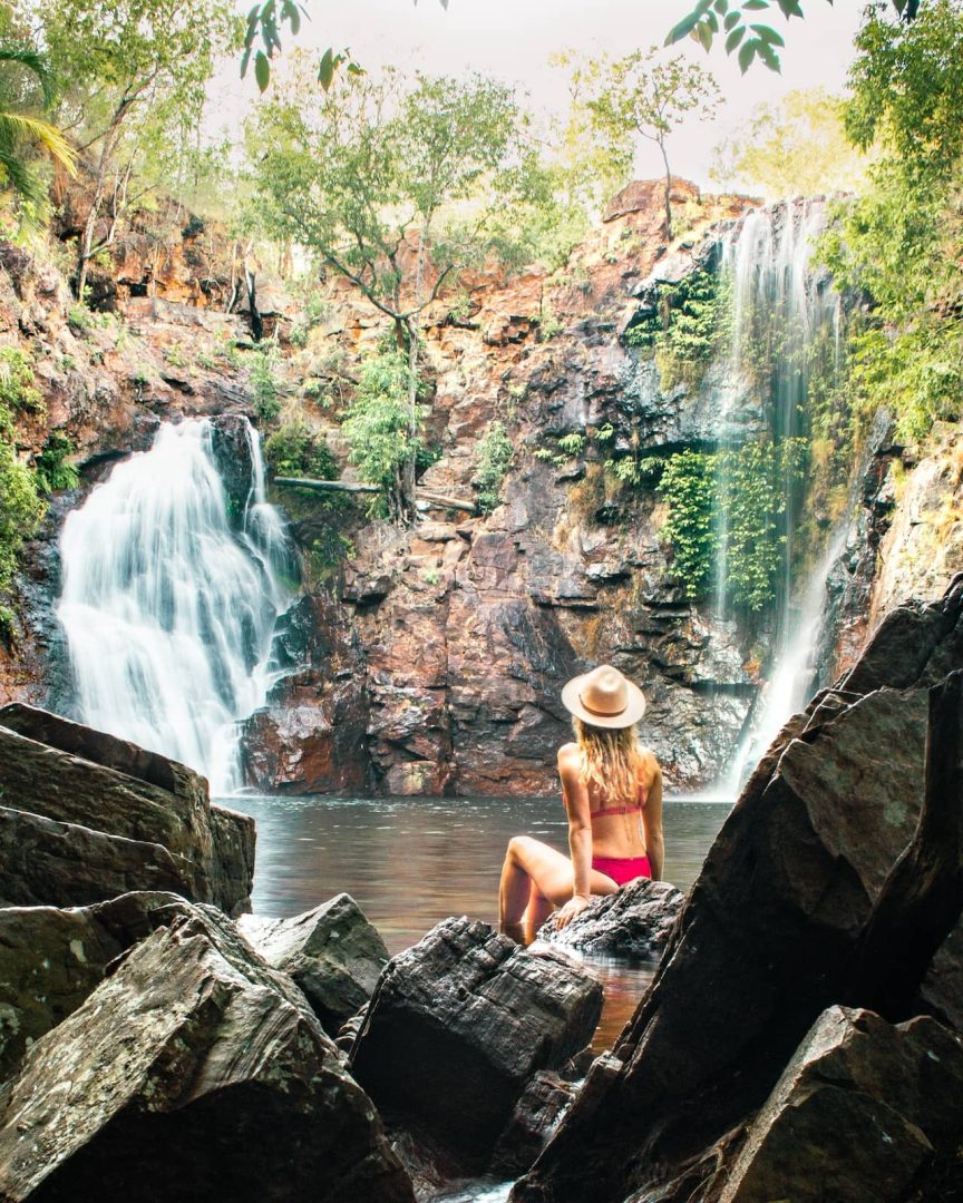 Woman in a red swimsuit and wide-brimmed hat sitting on rocks in front of Florence Falls cascading into a rock pool, captured on a Melbourne to Darwin road trip through the Top End.