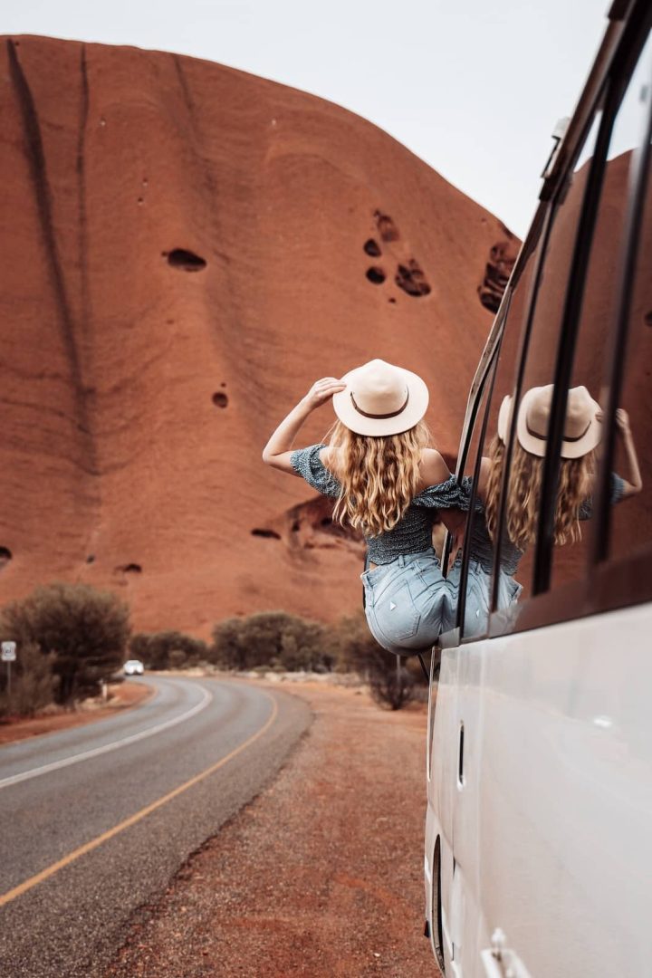 A woman in a wide-brim hat leans out of a white van window to admire Uluru up close during her red centre way adventure. The van curves along a quiet outback road with the towering sandstone behind her.