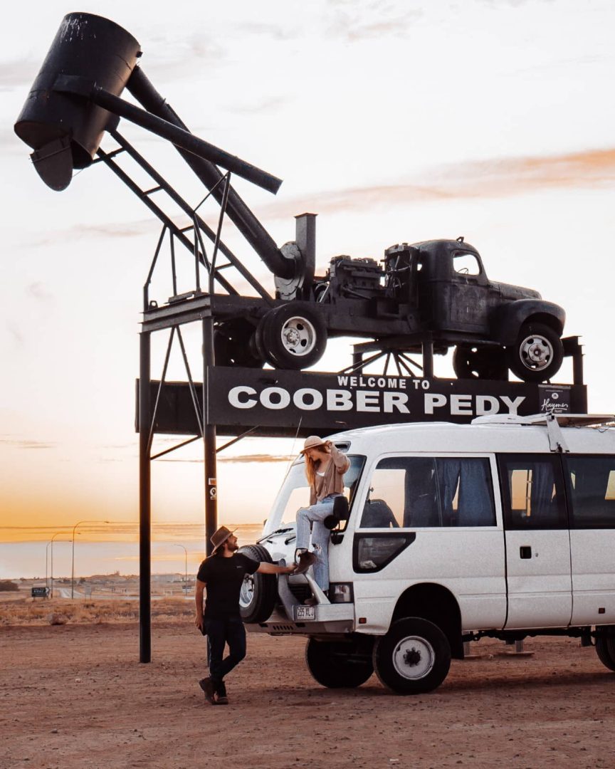 Couple with a van at sunset under the "Welcome to Coober Pedy" sign, featuring an old mining truck display, captured during a Melbourne to Darwin road trip through the Outback.
