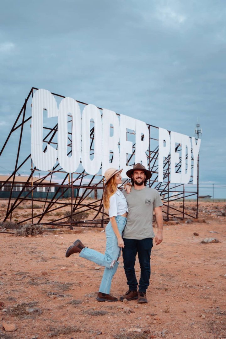 Smiling couple poses in front of the large white Coober Pedy sign in the red dirt Outback landscape, a classic photo stop on a Melbourne to Darwin road trip.