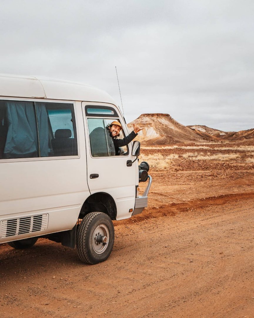 Man waves from a white van on a red dirt road with rugged hills in the background near Coober Pedy, showing life on the road during a Melbourne to Darwin road trip.