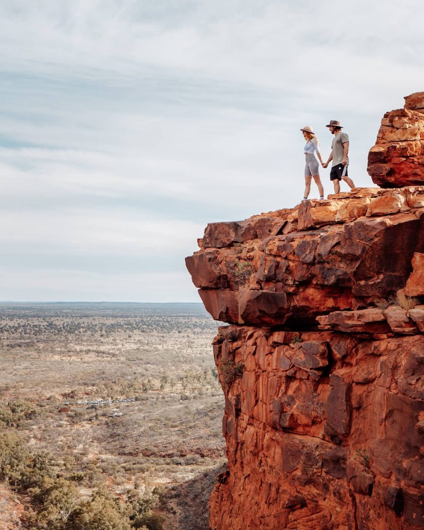 A couple stands on the edge of a towering red cliff at Kings Canyon holding hands, taking in the expansive outback views along the red centre way. The rocky ledge contrasts with the wide open landscape below.