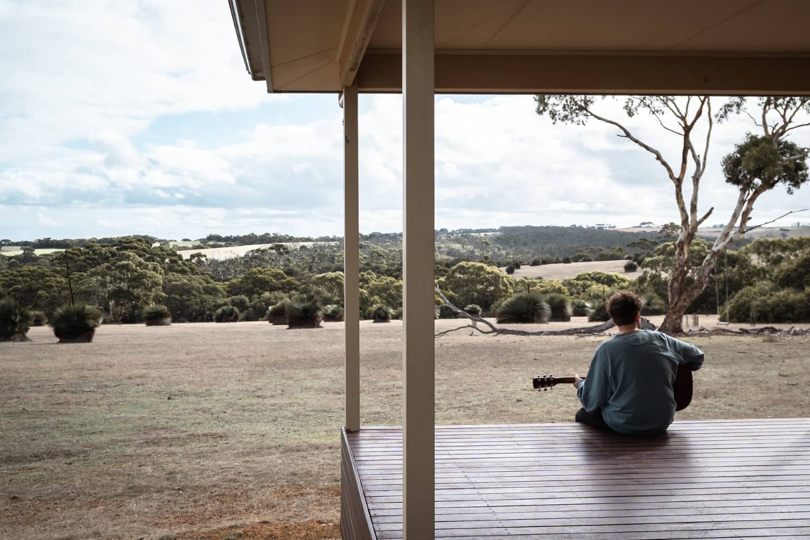 Man playing guitar on a cabin deck with wide views of farmland and native bush during a laidback kangaroo island road trip 3 days.