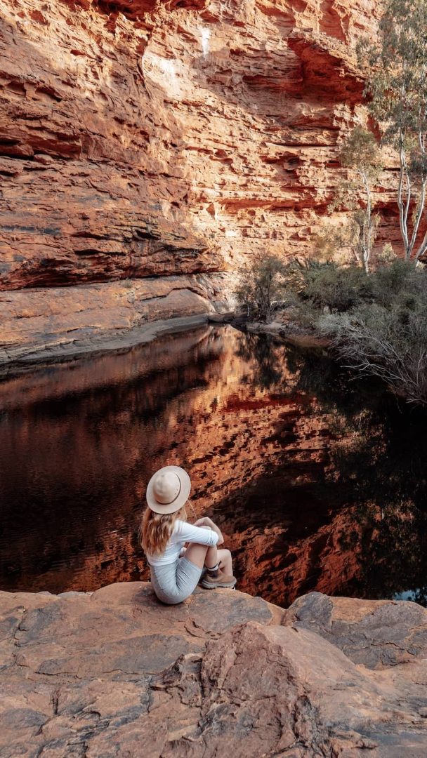 A woman sits by the Garden of Eden in Kings Canyon, a still waterhole surrounded by towering red canyon walls, a peaceful stop on the alice springs to uluru road trip. Reflections shimmer on the dark surface of the pool.