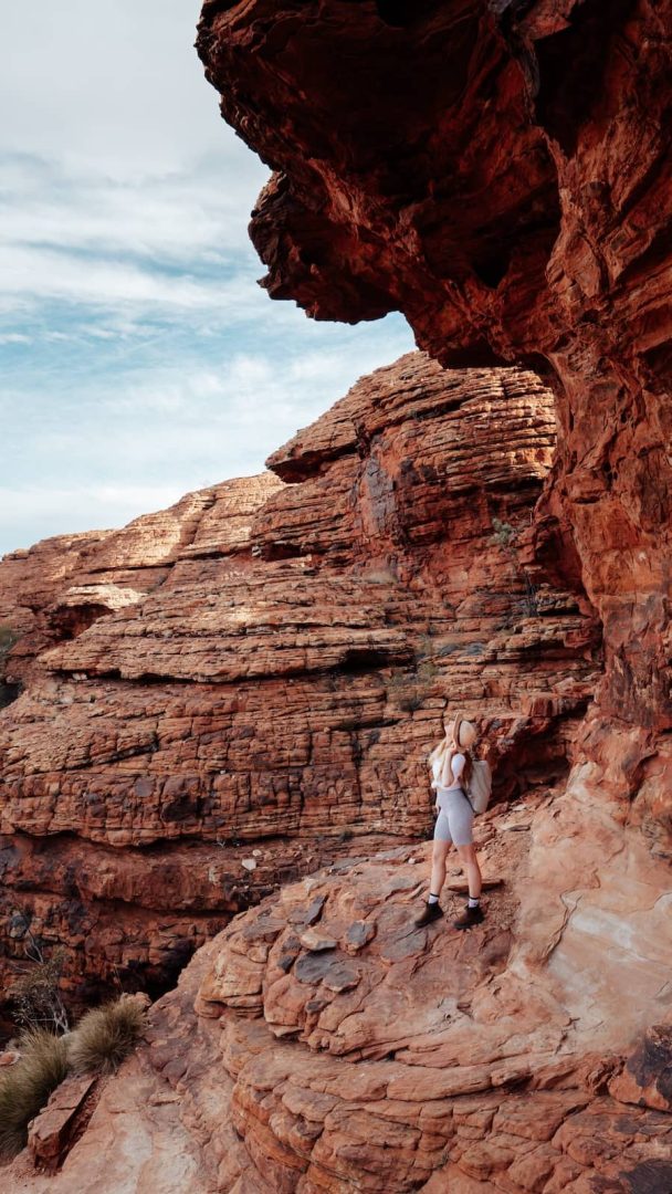 A hiker pauses beneath a jagged overhang while exploring the layered rock formations of Kings Canyon during a red centre way adventure. The rugged terrain showcases Central Australia's geological beauty.