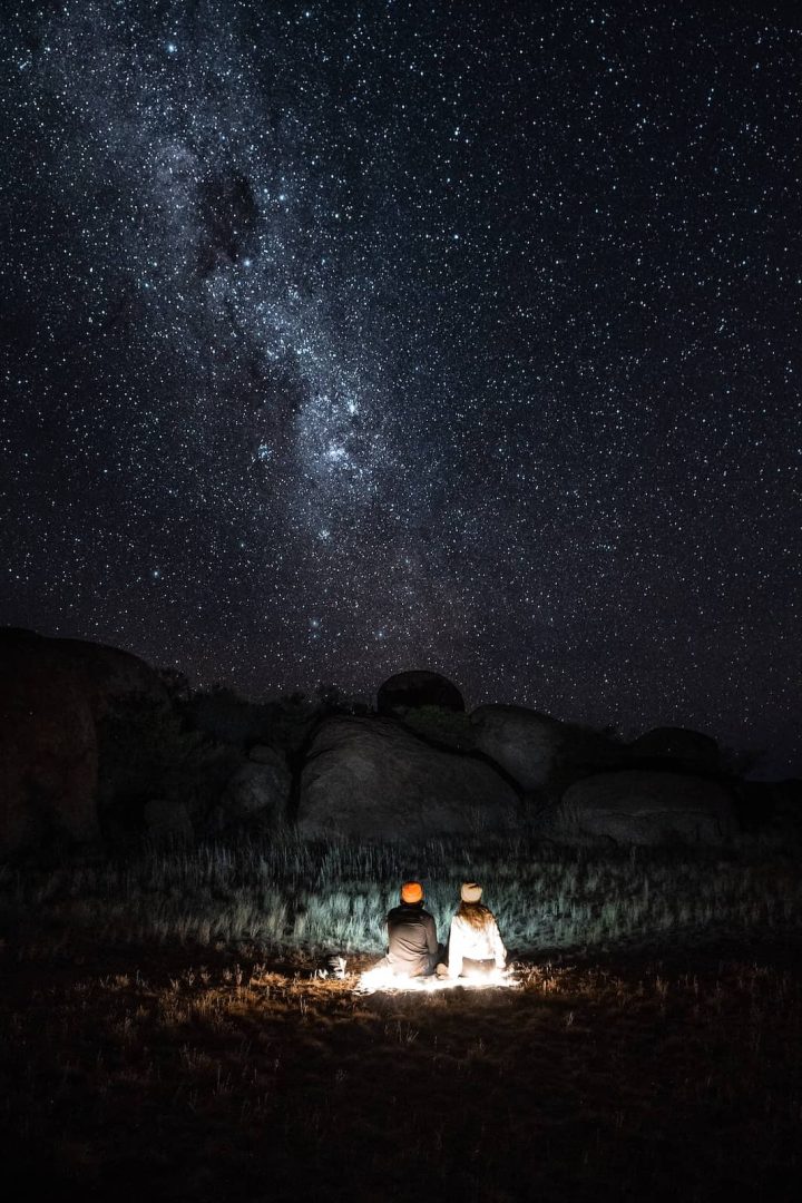 Couple sitting under the Milky Way with headlamps casting light around them at the Devils Marbles, enjoying stargazing on a Melbourne to Darwin road trip.