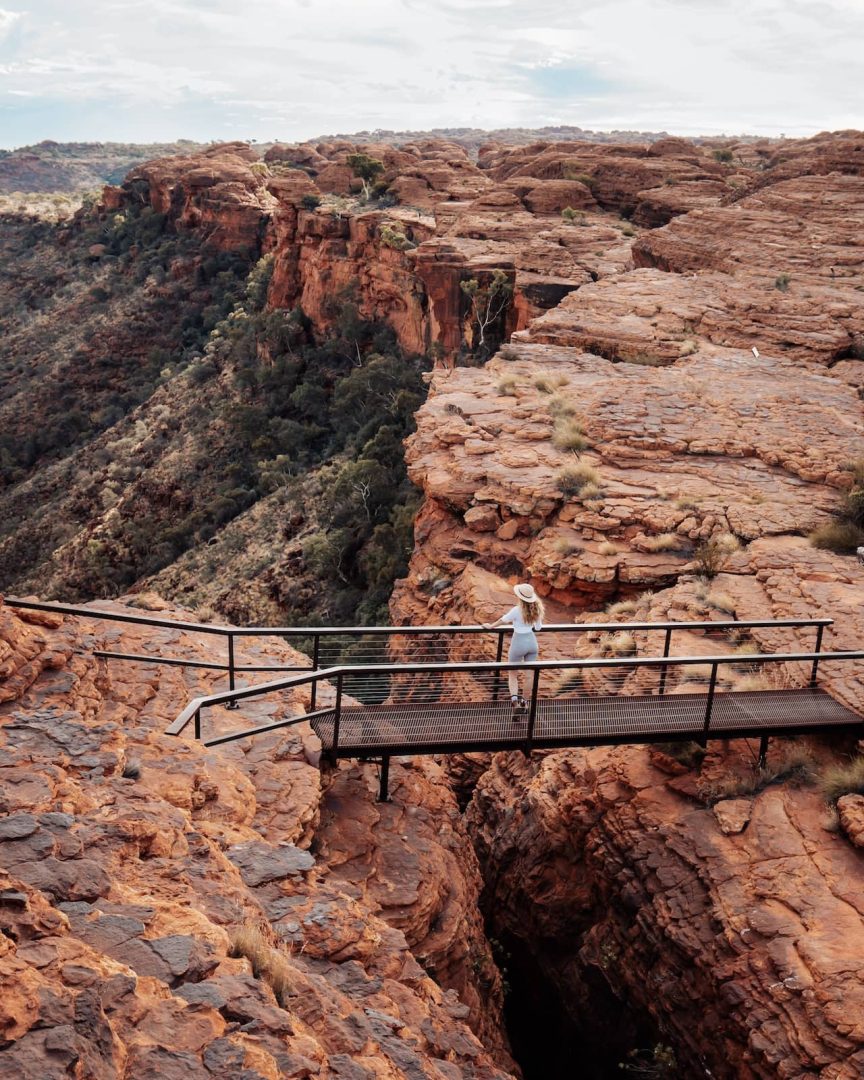 A woman walks across a metal viewing platform spanning a crevice at Kings Canyon, taking in the panoramic views of the rugged red landscape along the alice springs to uluru road trip.