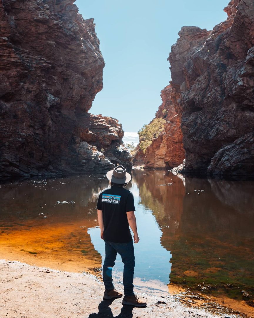A man in a hat stands at the edge of Ellery Creek Big Hole , surrounded by towering red cliffs during a stop on the red centre way. The still water reflects the rugged scenery.