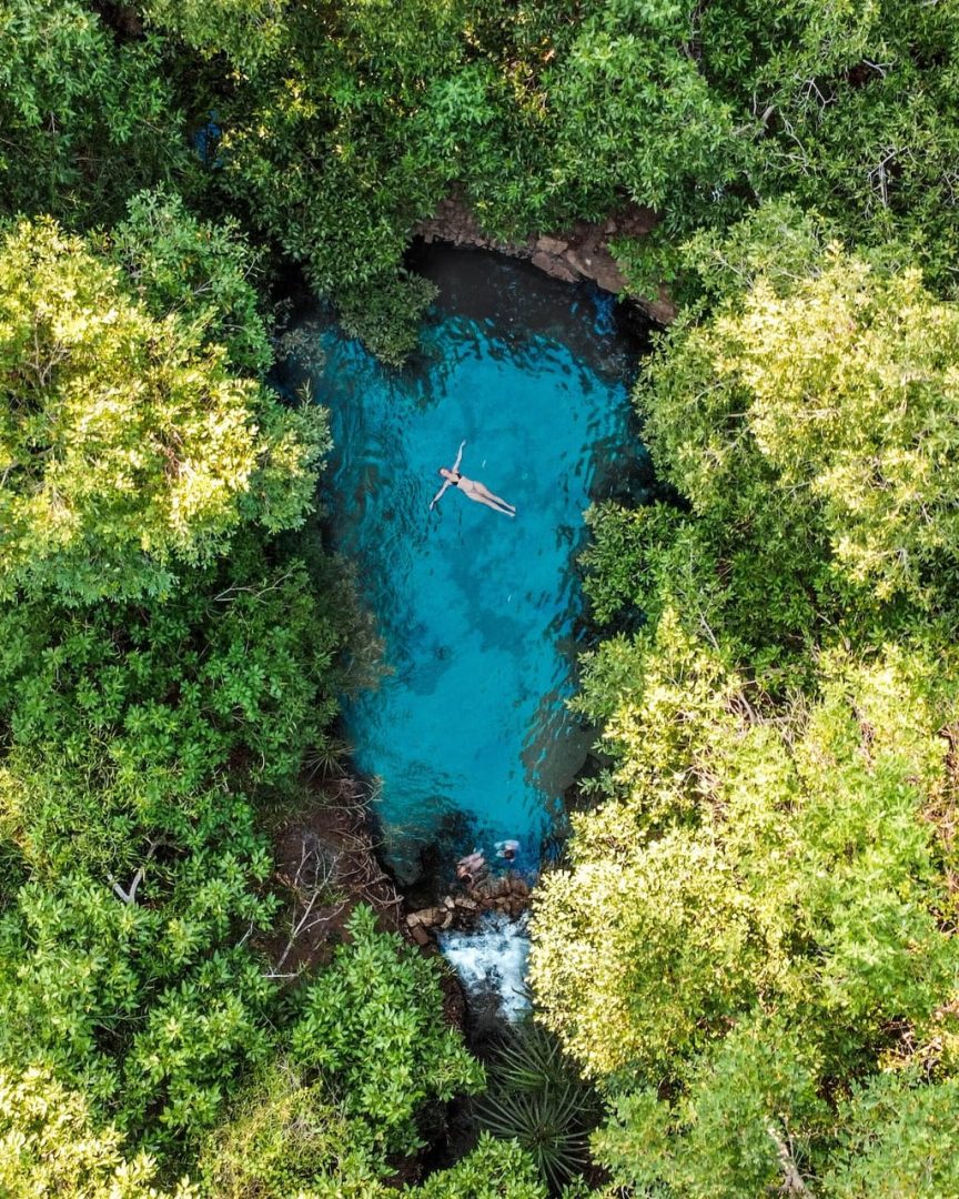 Aerial view of a person floating in the bright turquoise waters of the secluded Katherine Hot Springs, encircled by lush greenery, a tropical highlight of the Melbourne to Darwin road trip.