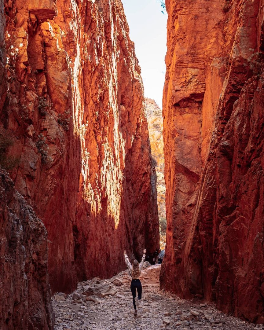 A woman walks between the steep red rock walls of Standley Chasm with arms raised in awe, a dramatic section of the red centre way near Alice Springs. The narrow chasm is filled with soft light.