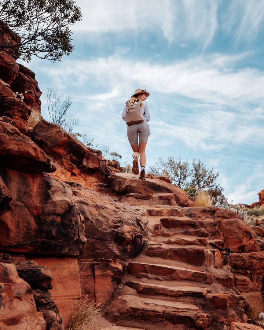 A woman with a backpack climbs carved stone steps through a rocky section of Kings Canyon, continuing her red centre way hike under a vivid blue sky. Wildflowers bloom beside the path.