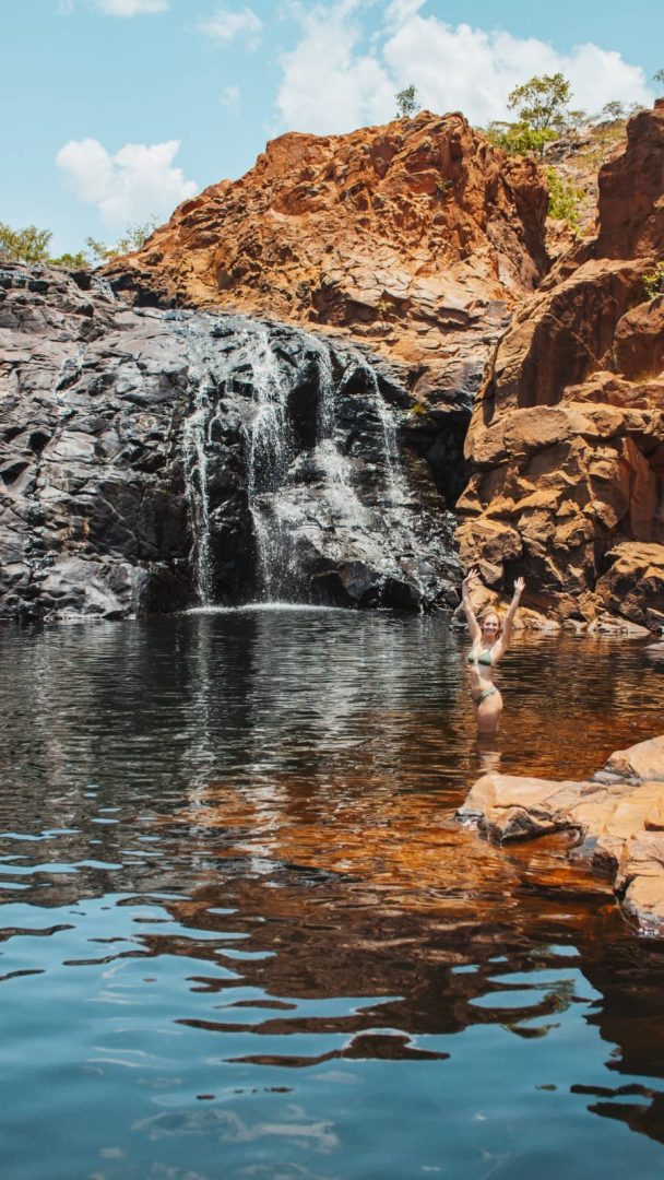 Woman standing in a rock pool beneath a small waterfall at Edith Falls, surrounded by red rocks, captured during a Melbourne to Darwin road trip.