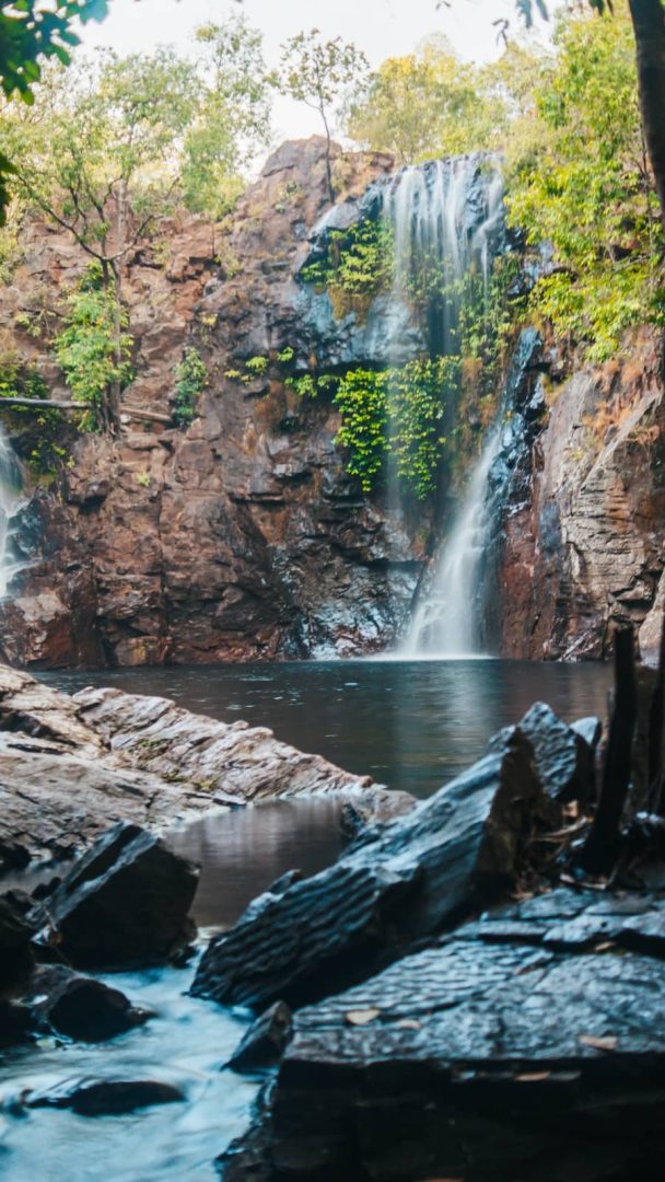 Twin streams of water cascade down mossy red rock walls into a quiet plunge pool at Florence Falls, a must-see waterfall on the Melbourne to Darwin road trip.
