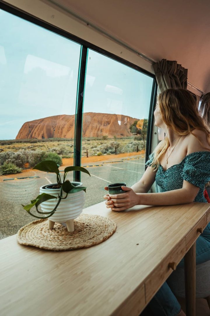 A woman holds a ceramic mug while gazing out the window of a campervan at Uluru, parked in the desert during an alice springs to uluru road trip. A potted plant and woven mat sit on the wooden table inside.