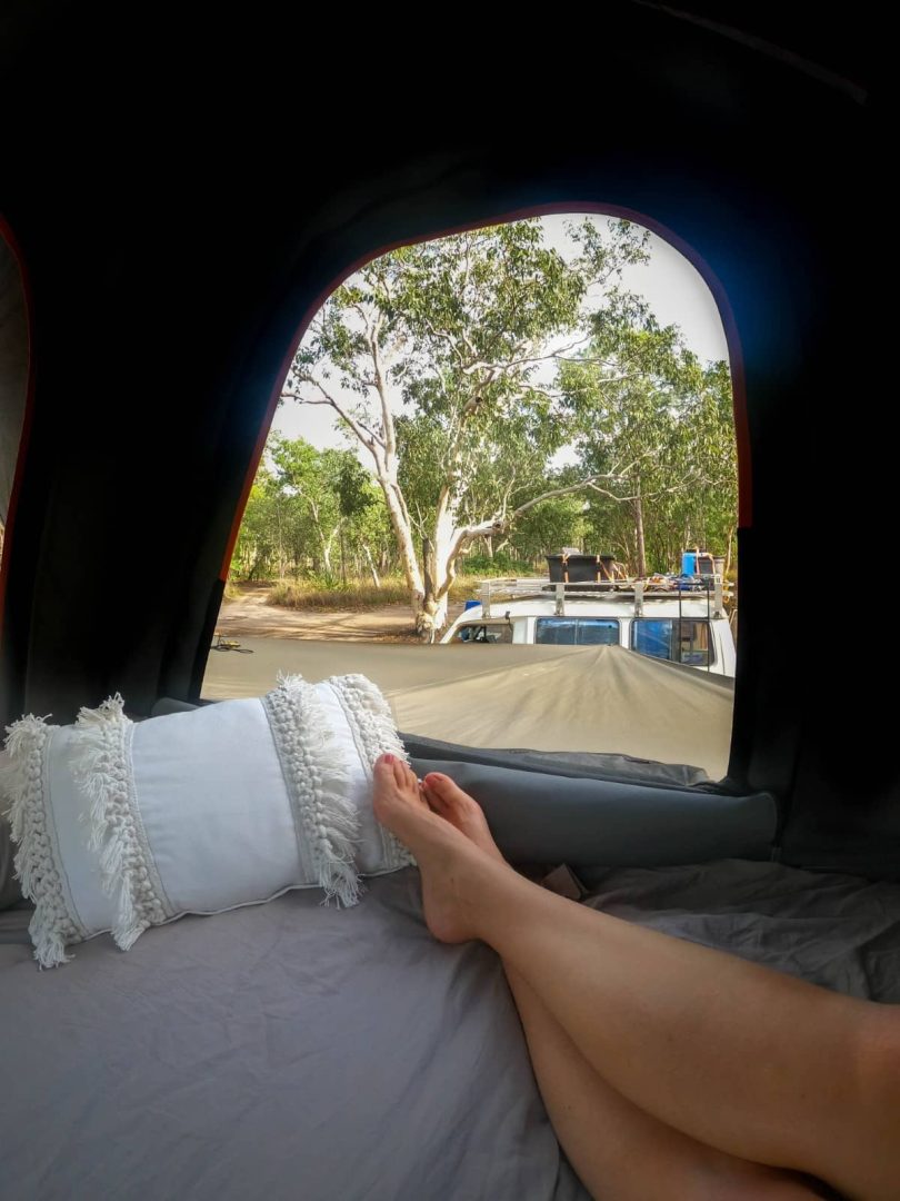 View from inside a rooftop tent looking out at trees and another camper setup in the bush, a peaceful overnight camp during a Melbourne to Darwin road trip.