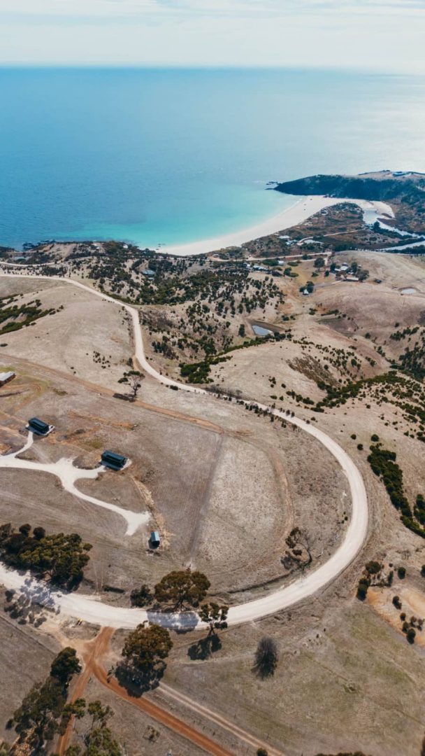 Aerial photo of winding unsealed roads through dry farmland leading to a stunning white-sand beach on a kangaroo island road trip 3 days.