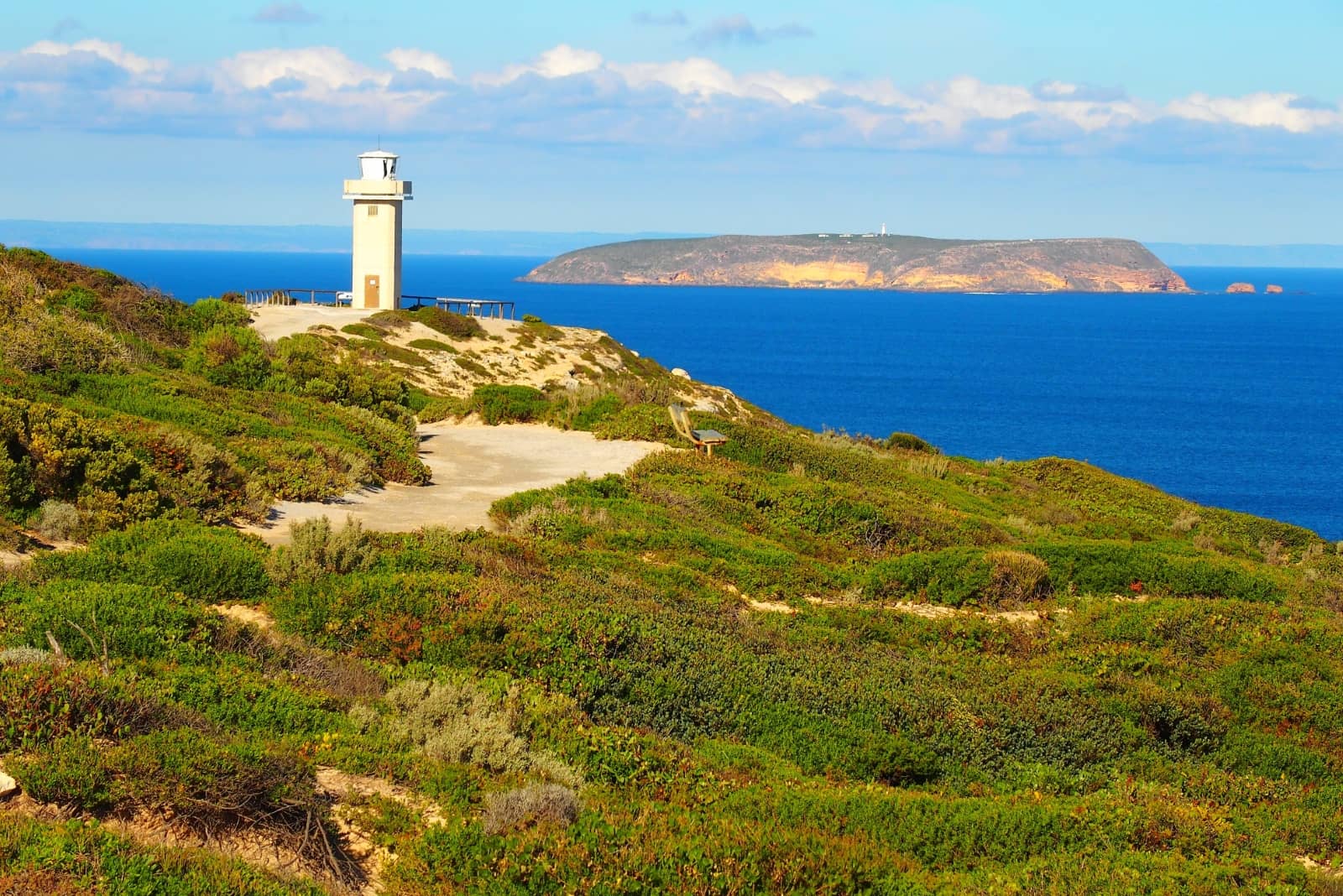 Cape Spencer lighthouse in Yorke Peninsula, south Australia. It's a lighthouse perched on a clifftop surrounded by green shrubs, overlooking deep blue ocean waters. A large island (Althorp Island) is visible across the water in the distance.