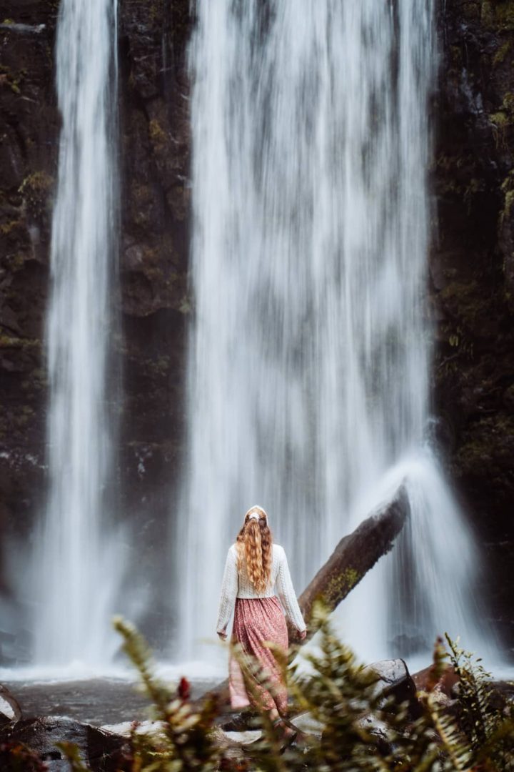 Woman with long hair in a pink skirt and jumper walking toward a the powerful Hopetoun Falls surrounded by mossy rock walls during a Melbourne to Darwin road trip.