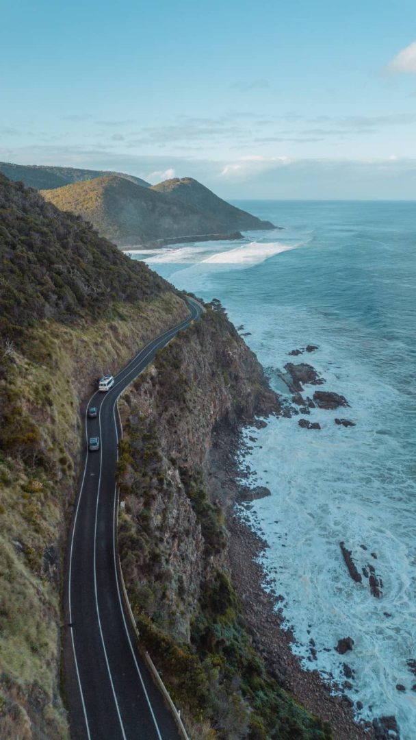 Cars drive along a winding cliffside road overlooking crashing ocean waves on the Great Ocean Road, an essential part of the Melbourne to Perth road trip.
