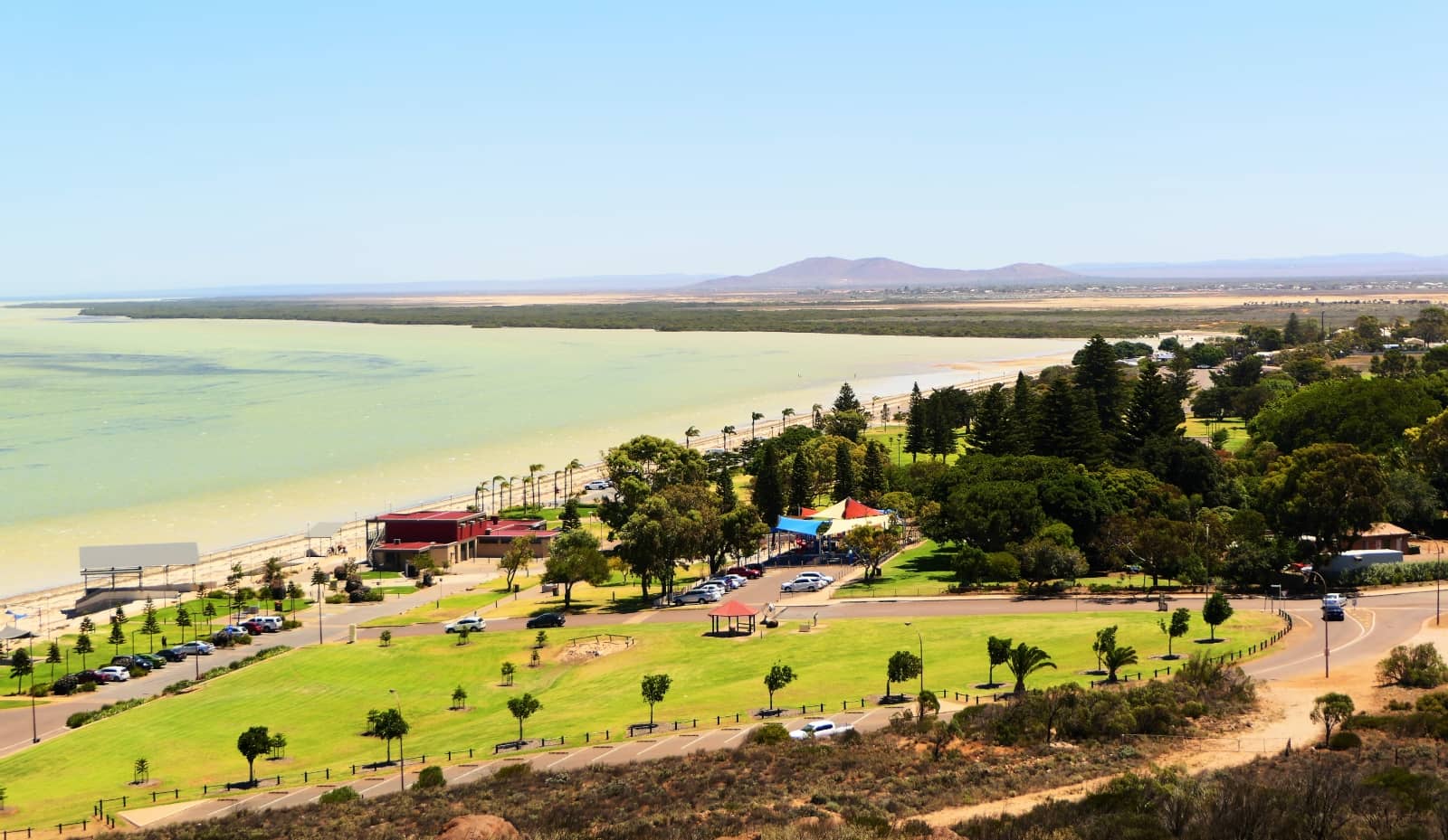 Aerial view of a coastal park and beachfront promenade in Whyalla, a town in South Australia, with green lawns, playgrounds, and palm-lined paths. Pale green water stretches along the coast with distant hills on the horizon.