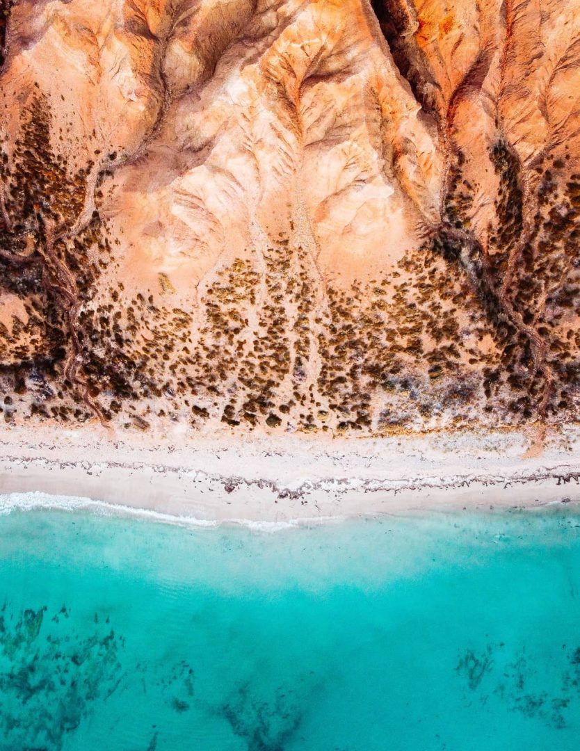 Aerial view of vibrant orange cliffs meeting clear aqua water on Sellicks Beach, near Adelaide, showcasing coastal beauty found on a Melbourne to Darwin road trip.