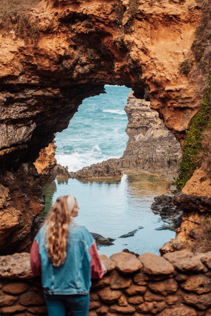 Woman looks through the natural rock formation known as The Grotto near Port Campbell, where the sea meets a tidal pool, a must-see stop on the Melbourne to Perth road trip.