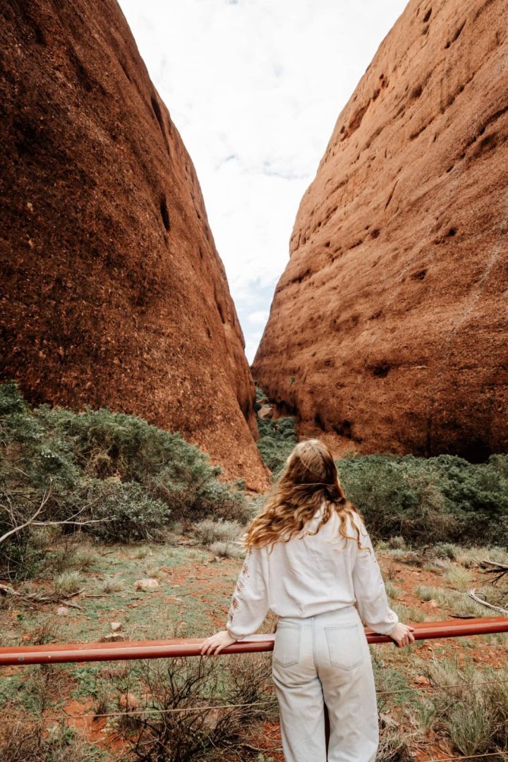 A woman stands at a lookout railing gazing into the narrow gorge between the domes of Kata Tjuta, one of the key sites along the red centre way. The red rock walls rise steeply on either side.