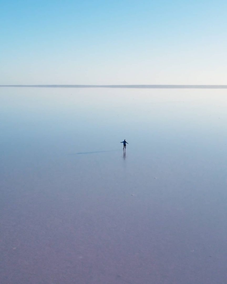 A person stands alone on the reflective pink surface of Lake Hart in South Australia under a soft blue sky, a surreal stop along the Melbourne to Darwin road trip