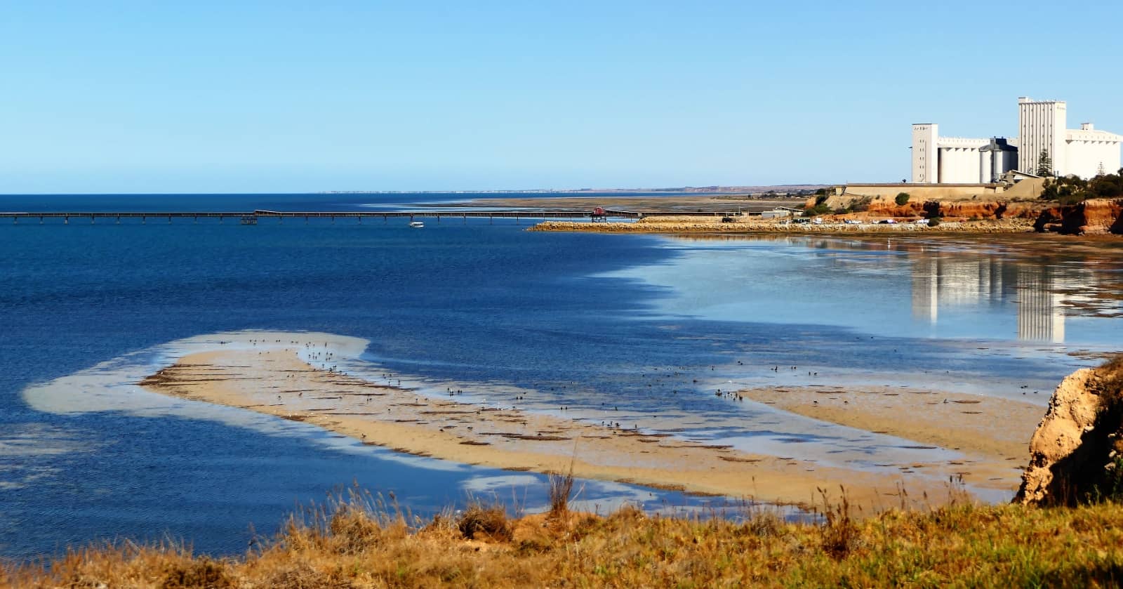 View of the Ardrossan coastline in South Australia with clear blue waters, a sandy tidal flat in the foreground, and a long jetty extending into the Gulf St Vincent. Large white grain silos sit prominently on the red cliffs in the background, reflecting in the calm shallows below.