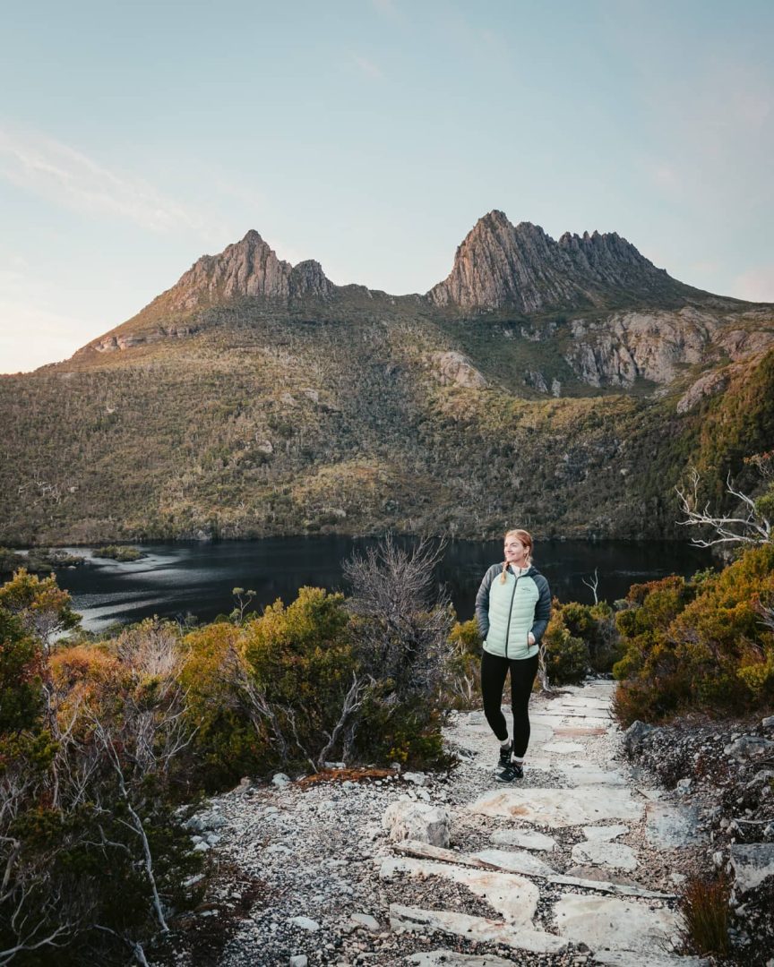 A woman hikes a rocky path overlooking Dove Lake with Cradle Mountain in the background, a key destination on a 7 day Tasmania itinerary.