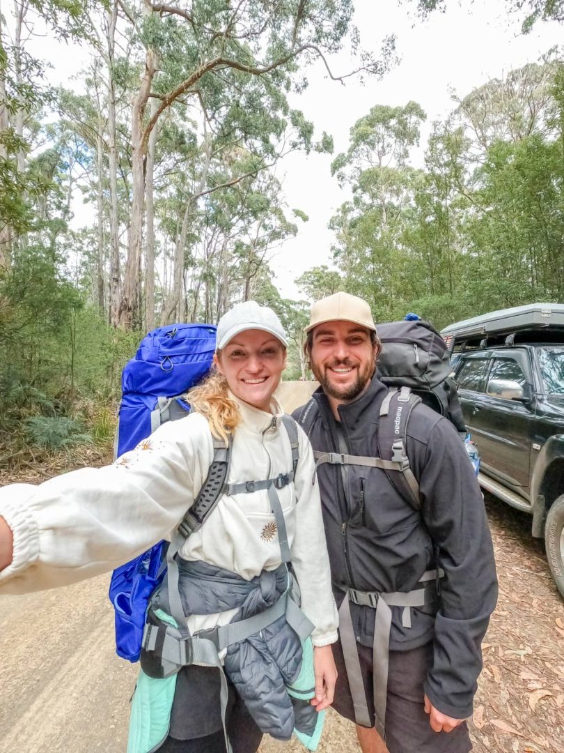 Smiling couple with hiking backpacks taking a selfie on a forest road, ready for an outdoor trek as part of their Tasmania Road Trip 10 Days.