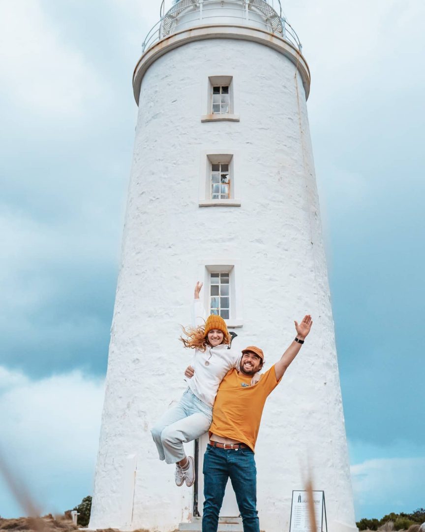 A happy couple pose in front of Cape Bruny Lighthouse under a stormy sky, one lifting the other in celebration during their great eastern drive.