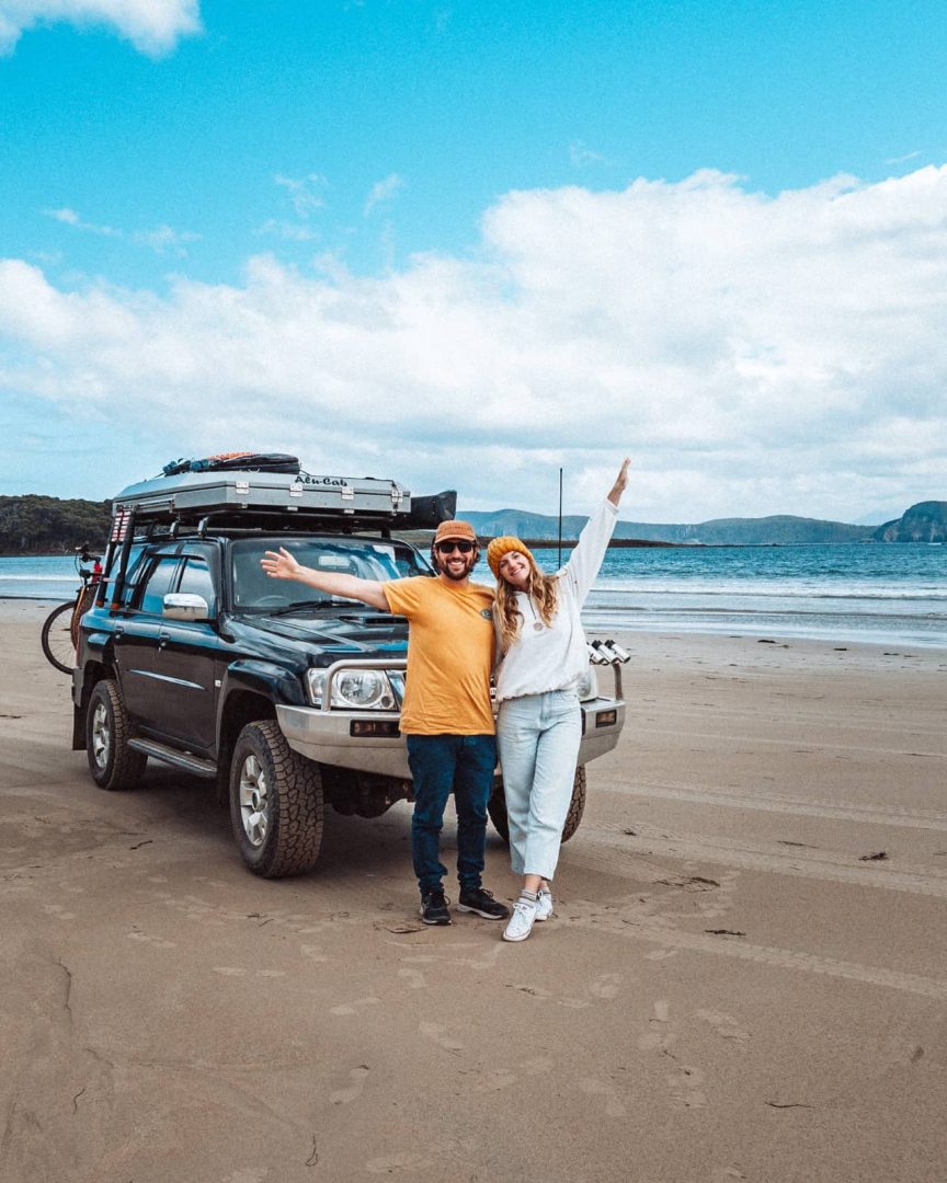 Smiling couple poses in front of a 4WD camper setup on a Tasmanian beach, capturing the freedom of a 7 day Tasmania road trip adventure.