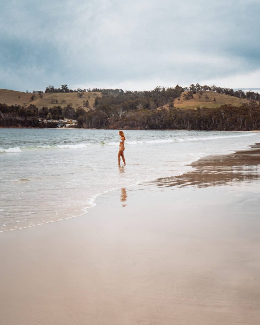 Woman standing in shallow waves at Binalong Bay, surrounded by calm waters and distant hills under an overcast sky, part of a coastal stop on a Tasmania Road Trip 10 Days.