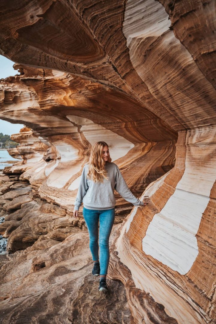 Woman walking beside the colourful layered rock formations of the Painted Cliffs on Maria Island, a geological highlight on a 10 day Tasmania road trip.
