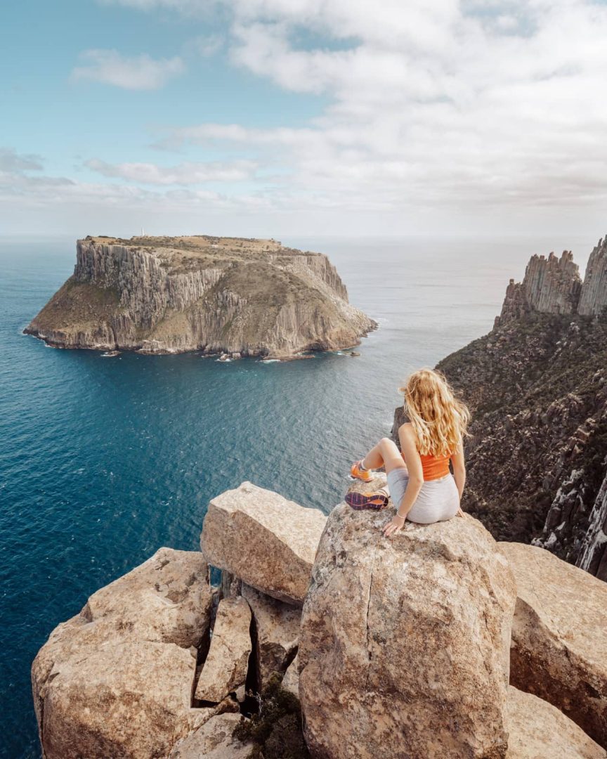 A woman sits on a rocky cliff from the Three Capes Track, overlooking the dramatic seascape and Tasman Island, part of a scenic stop on a 7 day Tasmania itinerary.