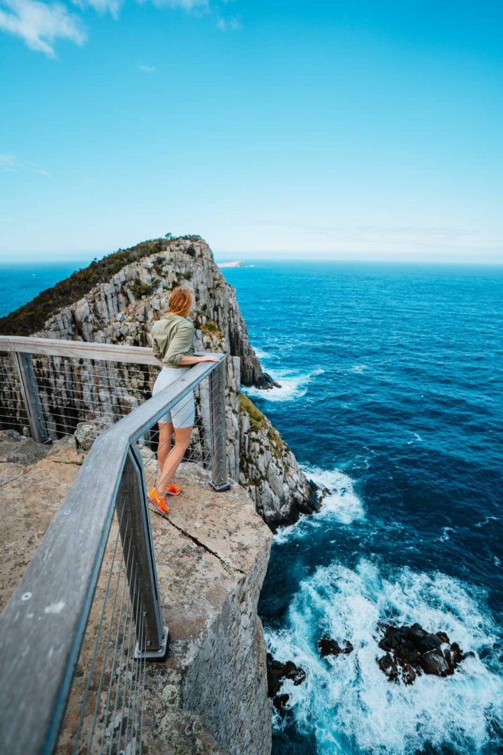 Woman standing on a fenced clifftop lookout gazing over rugged sea cliffs and crashing waves at Cape Hauy, featured on a 7 day Tasmania road trip.