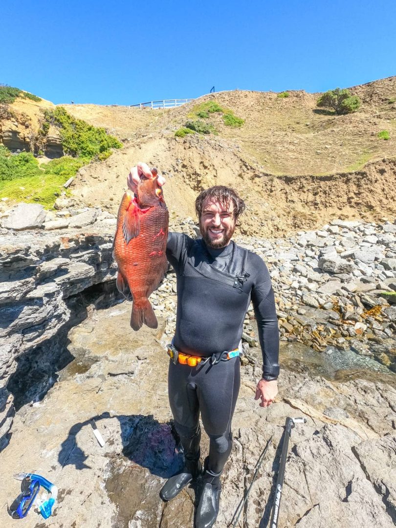 Man in wetsuit proudly holding a large fish beside rocky shoreline, after spearfishing off the east coast during a Tasmania Road Trip 10 Days adventure.