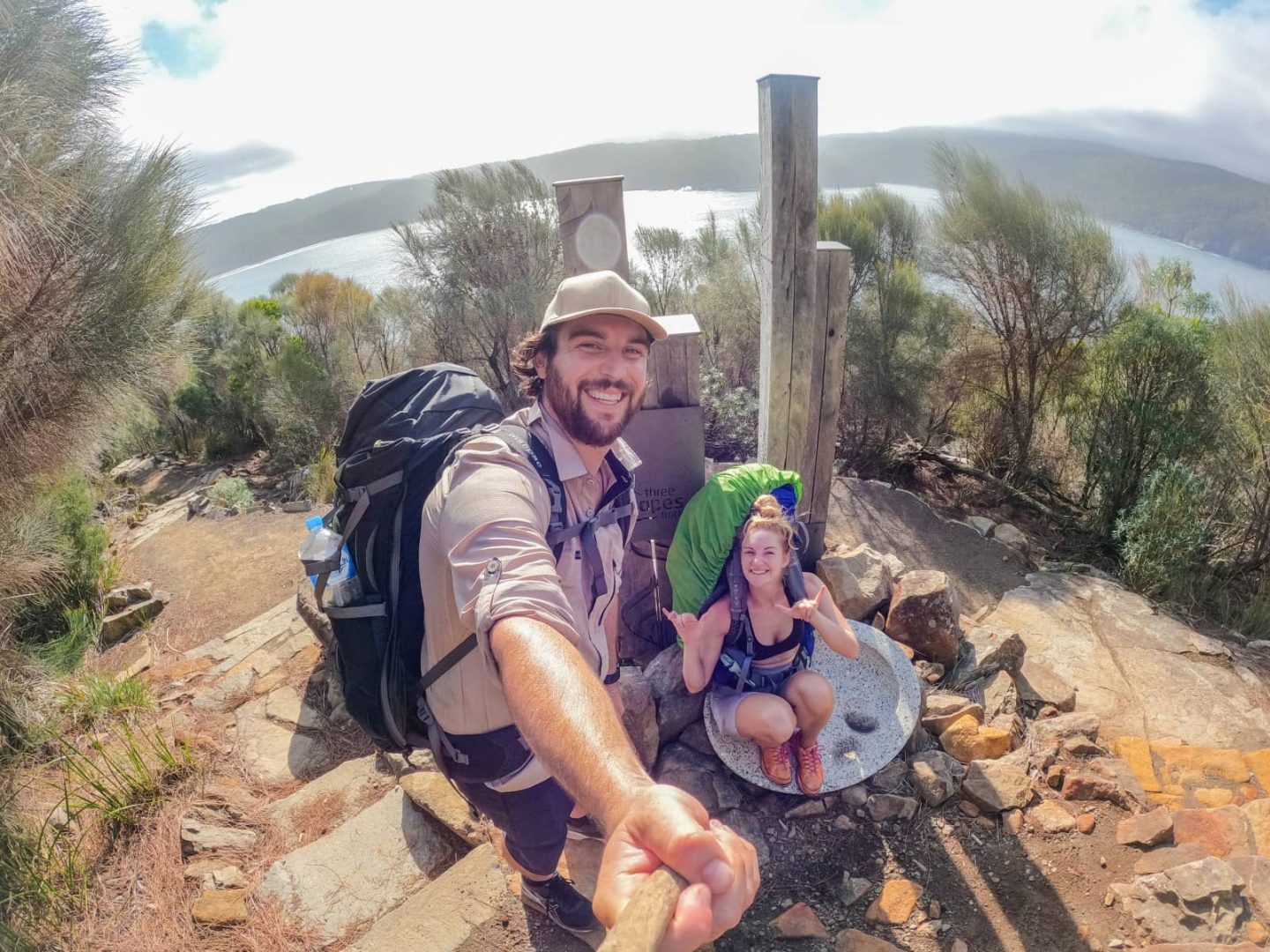 Two hikers smiling at a scenic lookout with backpacks on the Three Capes Track, a highlight of the Tasmania East Coast Road Trip.