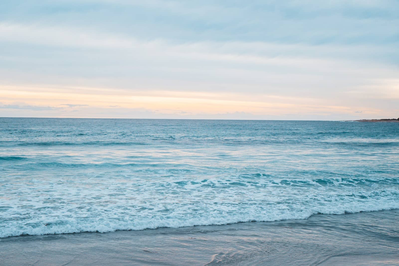 Soft waves roll onto a peaceful beach under pastel skies at sunset, capturing the tranquil coastal beauty found on a 7 day Tasmania road trip.