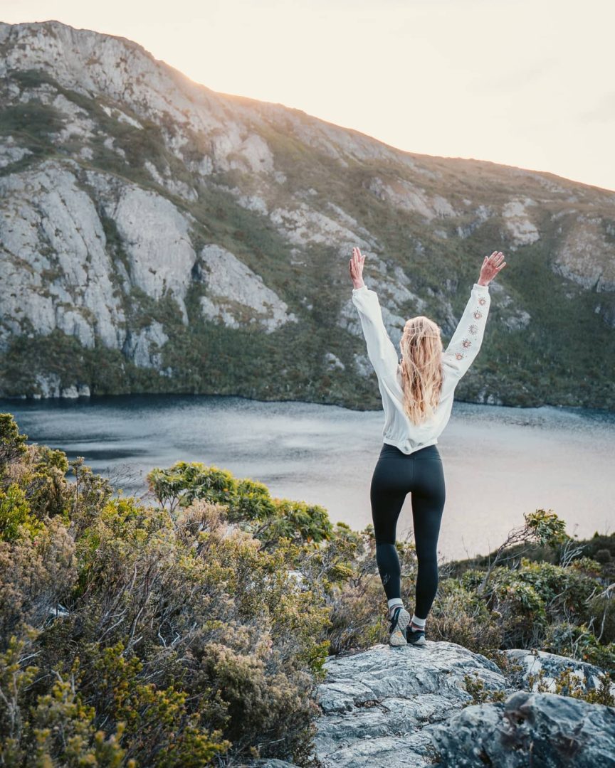 Woman raising arms while overlooking a lake beneath Cradle Mountain at sunset, celebrating a scenic moment on a Tasmania itinerary 10 days.