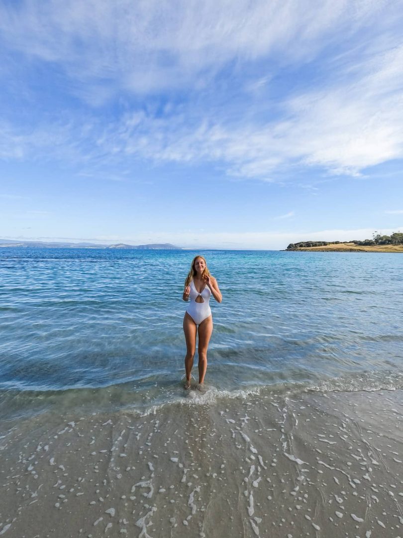 Woman in a white swimsuit standing ankle-deep in crystal-clear ocean water at a quiet beach stop on the Tasmania East Coast Road Trip.